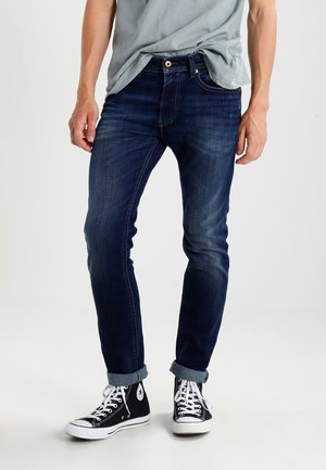 Personne portant un jean slim bleu foncé retroussé et des baskets montantes noires avec des lacets blancs, debout devant un fond uni.