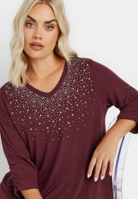 Burgundy pitkähihaisessa paidassa on V-mallinen kaulus, ja siinä on hopeinen kivikoriste, joka tiheästi alkaa rinnasta ja häipyy harvinaisemmaksi.