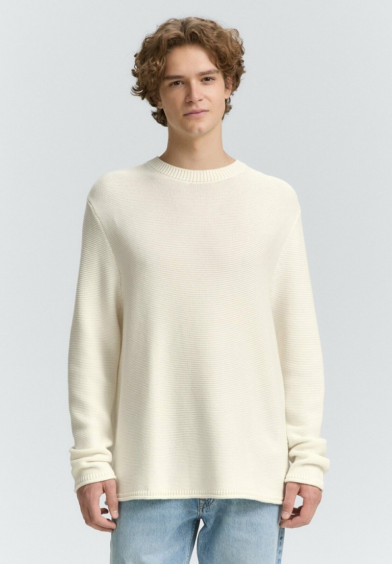 Jongen met krullend haar die een losse, crèmekleurige gebreide trui en lichtblauwe jeans draagt, staand tegen een neutrale achtergrond.