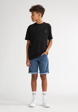 Zwart katoenen T-shirt met een klein logo, gecombineerd met spijkershorts met opgerolde pijpen. Witte sneakers en gestreepte sokken maken de outfit compleet.