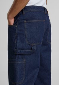 Pantalons en denim bleu foncé avec une coupe décontractée, des poches arrière et des poches latérales supplémentaires. Détails avec des coutures orange contrastantes.