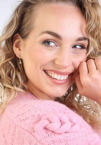 Roze gebreide trui met een gestructureerd ontwerp en gevlochten accent. Gouden ringoorbellen met heldere stenen. Kruidig haar los gestyled.