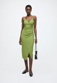 Zaļas pieguļošas kleitas ar plānām lencītēm, drapējumu dizainu un sānu šķēlumu. Modele tur melnu rokassomu un valkā melnas kurpes ar kvadrātveida degunu.