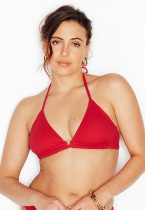 Femme aux cheveux bruns portant des boucles d'oreilles créoles dorées, un haut de bikini rouge à col halter, et un bracelet doré sur un fond blanc.