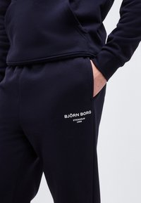 Κοντινό πλάνο ενός ατόμου που φοράει μαύρο παντελόνι φόρμας και φούτερ, με το χέρι στην τσέπη, και λευκό λογότυπο Björn Borg Stockholm 1956 στα παντελόνια.