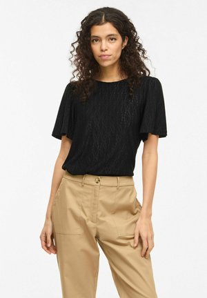 Kvinde med krøllet hår iført sort kortærmet top med tekstureret mønster og beige bukser med høj talje, stående foran en hvid baggrund.