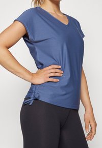 Mujer vestida con una blusa azul de manga corta y escote en V, con un detalle de lazo lateral y leggings negros, posando con una mano en la cadera.
