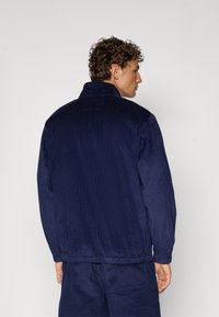 Marine kordfløyelsjakke med høy krage, elastisk livlinje og ribbestruktur. Har en sømløs ryggdesign med en subtil baksløyfe detalj.