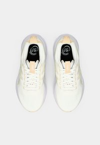 Par de zapatillas de correr blancas y color melocotón claro con cordones, vista superior, mostrando plantillas negras etiquetadas con "+10.3 kg CO2" huella de carbono.