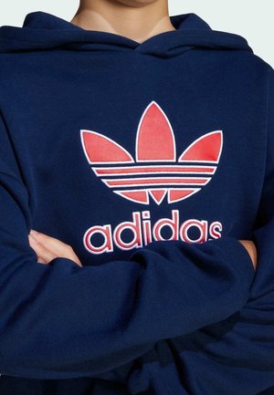 Person wearing navy blue hoodie with red and white Adidas Trefoil logo and the word "adidas" on the chest, arms crossed.