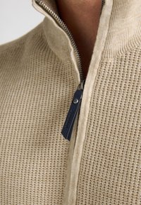 Beige stickad tröja med dragkedjekrage, prydd med ett texturerat mönster och en marinblå dragkedja med logotypdetalj.