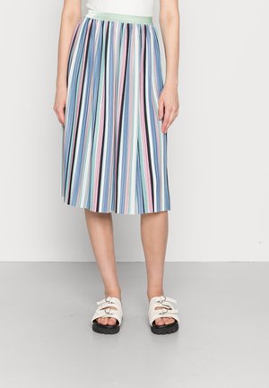 Vrouw draagt een geplooide midi-rok met verticale pastelkleurige strepen en witte sandalen met dubbele gespsluitingen, staand op een grijze vloer.