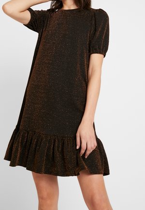 Mujer vistiendo un vestido negro de manga corta hasta la rodilla con brillo cobre y dobladillo con volantes, de pie frente a un fondo liso.