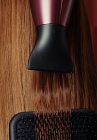 Hårtork med metallisk rosa och svart design som blåser luft på slätt, rakt, långt brunt hår, tillsammans med en svart borste med texturerade borst.