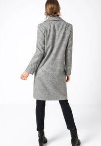Grå rutig kappa med lång siluett, enkelknäppt design och slag. Strukturerad tyg med uttalade rutor.