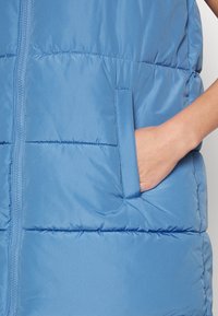 Ljusblå, ärmlös puffväst i glänsande tyg. Har en blixdragkedja framtill och en sidoficka med platt söm och diskreta sömnadsdetaljer.