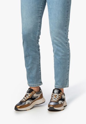 Persona che indossa jeans blu e sneakers multicolori con dettagli neri, marroni, bianchi e in rete, in piedi su uno sfondo bianco.