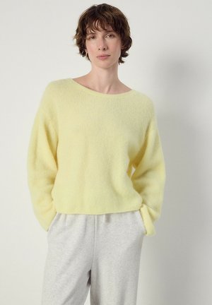 Frau mit kurzen braunen Haaren, die einen weichen gelben Pullover und hellgraue Hosen trägt und vor einem einfarbigen weißen Hintergrund steht.