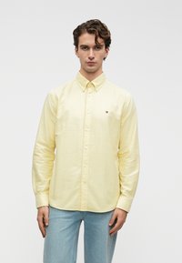 Ung mand iført en lys gul skjorte med lange ærmer og lyseblå jeans, stående mod en ensfarvet hvid baggrund.