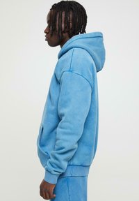 Haut à capuche bleu clair avec poches kangourou, poignets côtelés et capuche à cordon, fabriqué en tissu doux et texturé.