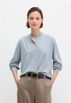 Kobieta o krótkich brązowych włosach, ubrana w jasnoniebieską koszulę na guziki, beżowe spodnie i czarny pasek, stoi z rękami w kieszeniach.