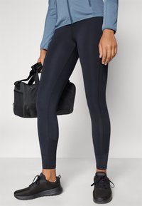 Άτομο που φοράει μαύρα κολάν, μαύρα αθλητικά παπούτσια και μπλε μπουφάν, κρατώντας μια μαύρη τσάντα ταξιδιού στο πλάι του.