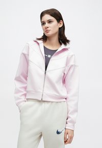 Νεαρή γυναίκα ντυμένη με έναν ανοιχτό ροζ φερμουάρ από μπουφάν πάνω από ένα σκούρο τοπ και με μπεζ φόρμες που έχουν μπλε λογότυπο, στέκεται μπροστά σε λευκό φόντο.