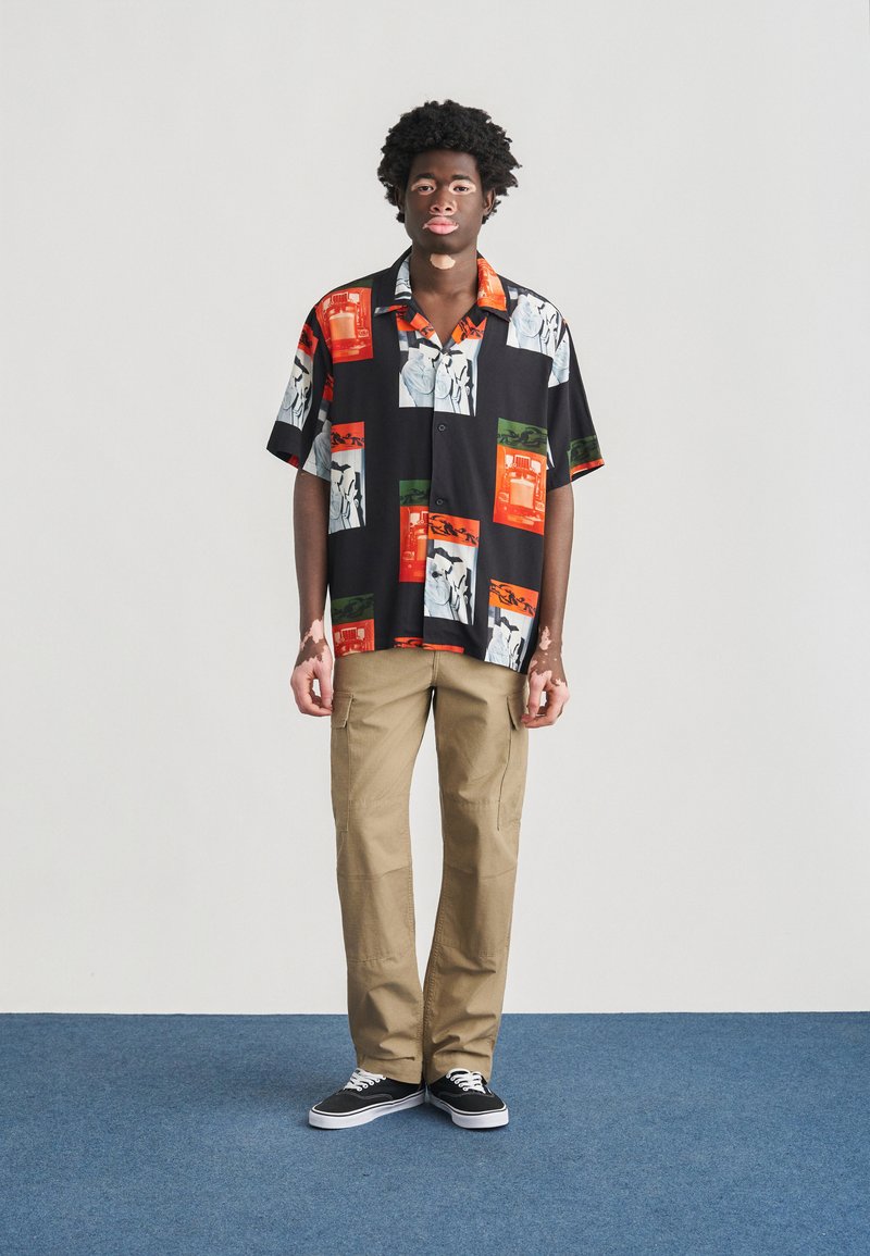 Maglietta nera a maniche corte con stampe grafiche colorate, abbinata a pantaloni cargo beige e sneaker nere. Tessuto liscio con vestibilità rilassata.