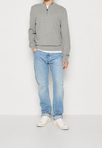 Person klädd i en grå kvartdragningströja, ljusblå jeans med uppvikningar, vit t-shirt och vita sneakers med beige logotyper, stående med en hand i fickan.