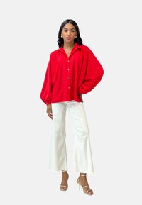 Red oversized button-up shirt with balloon sleeves, paired with white wide-leg pants and gold heeled sandals. Smooth texture, glossy finish.