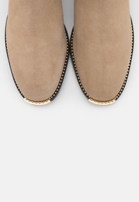 Botas de tobillo de ante beige con punteras redondeadas, suelas negras y acentos de metal dorado a lo largo del borde, con una textura suave.