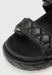 Sandales à plateforme noires avec texture matelassée, dotées d'une large sangle et d'accents en métal doré, conçues avec une semelle intérieure rembourrée.