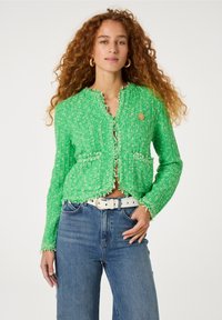 Groene, getextureerde cardigan met een franjeszoom, voorzien van een ronde hals, voorzakken en een gouden accentknop. Draagt bij hoge taille jeans.