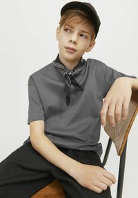 T-shirt grigio a maniche corte abbinato a pantaloni neri. Una bandana nera con motivi bianchi legata intorno al collo e un cappellino nero indossato all'indietro.