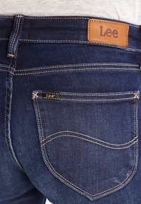 Vaqueros de mezclilla azul oscuro con un parche de cuero con el logo de Lee en la cintura y costuras amarillas en contraste. Presenta un bolsillo trasero visible con una etiqueta de logo.