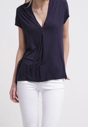 Vrouw draagt een marineblauw V-hals topje met korte mouwen en plooidetail aan de voorkant, gecombineerd met witte nauwsluitende broek tegen een effen achtergrond.