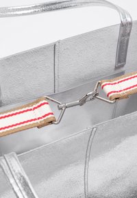 Srebrna torebka tote o fakturze z metalicznym połyskiem z odpinanym kremowym paskiem z czerwonymi i beżowymi paskami, zabezpieczonym metalowym zapięciem wewnątrz torby.