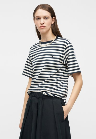 Γυναίκα που φοράει μπλε σκούρο και λευκό ριγωτό μπλουζάκι, κολιέ με μαργαριτάρια και ψηλόμεση μαύρη πλισέ φούστα, στέκεται με το ένα χέρι στην τσέπη.