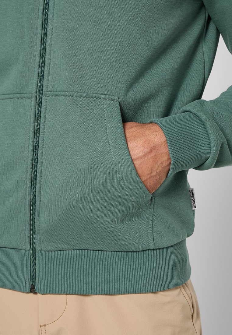 Zielona bluza z kapturem na zamek, z prążkowanymi mankietami i dołem, bocznymi kieszeniami oraz fakturowanym materiałem. Wyświetl pokazuje tylko górną część ciała i ręce.