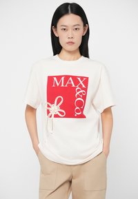 Γυναίκα με ίσια μαύρα μαλλιά, ντυμένη με λευκό T-shirt που έχει κόκκινο λογότυπο «MAX&Co.» και μπεζ παντελόνι, με τα χέρια στις τσέπες.