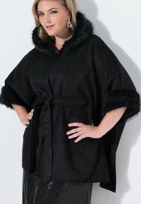 Capa negra con borde de piel sintética y capucha, que presenta un cinturón de lazo en la cintura y mangas anchas. Textura suave con acentos brillantes.