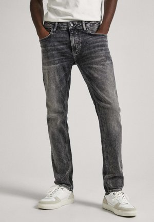 Mężczyzna w wyblakłych czarnych obcisłych dżinsach z rękami w kieszeniach i białych trampkach, stojący na jasnym, jednolitym tle.