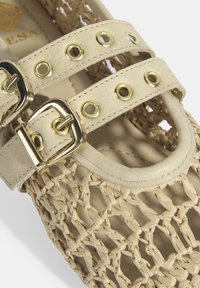 Beige gewebte Sandale mit Lederriemen, ausgestattet mit goldenen Beschlägen und mehreren Ösen. Offenes Design mit strukturierter Oberfläche.