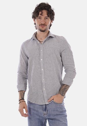 Uomo con capelli ricci che indossa una camicia a righe con bottoni e jeans, in piedi con una mano in tasca, mostrando tatuaggi sugli avambracci.