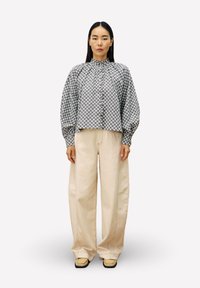 Frau mit langen schwarzen Haaren, die eine gemusterte blau-graue Bluse, breite beige Hosen und beige Schuhe trägt, steht vor einem schlichten hellen Hintergrund.