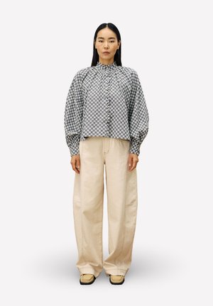 Femme aux longs cheveux noirs portant un blouson à motifs bleu-gris, un pantalon large beige et des chaussures beiges, se tenant devant un fond clair.