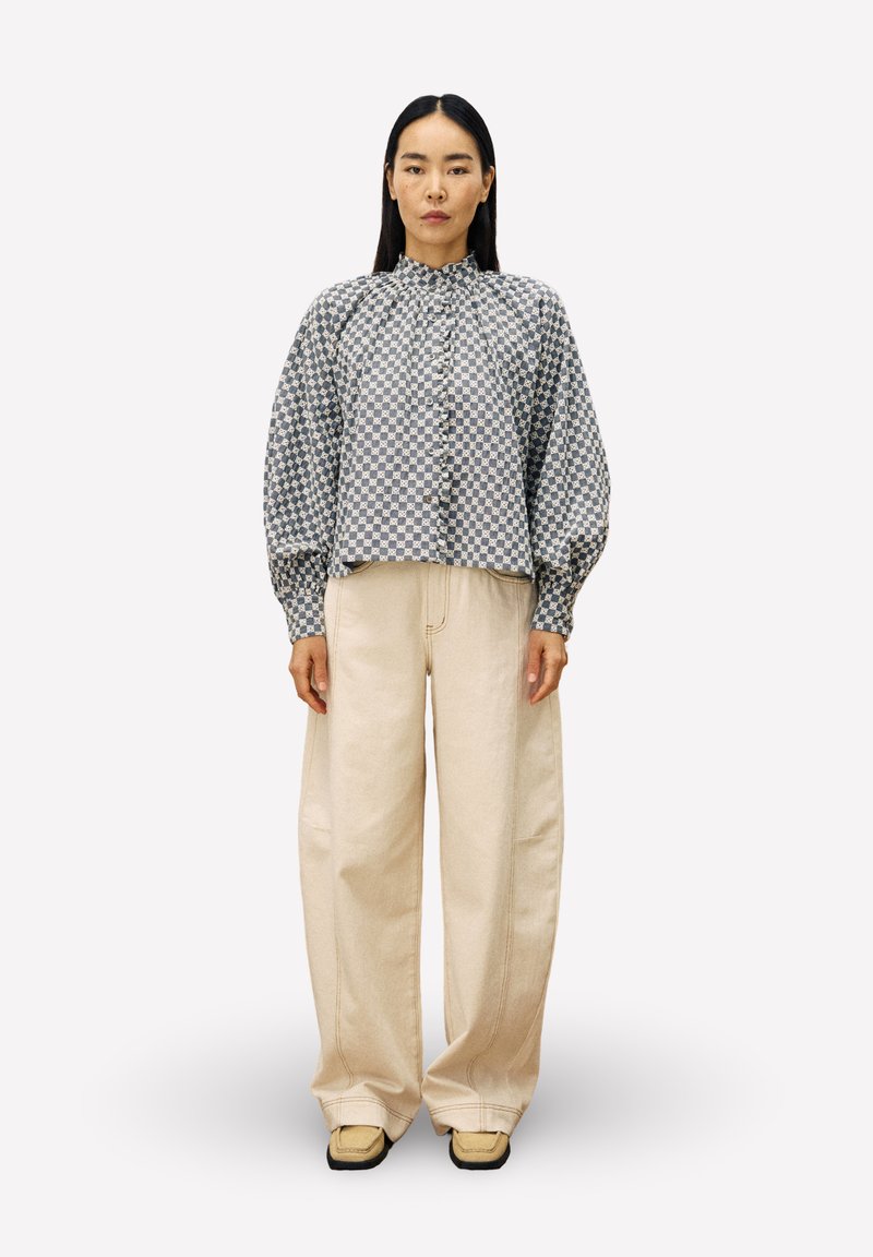 Frau mit langen schwarzen Haaren, die eine gemusterte blau-graue Bluse, breite beige Hosen und beige Schuhe trägt, steht vor einem schlichten hellen Hintergrund.