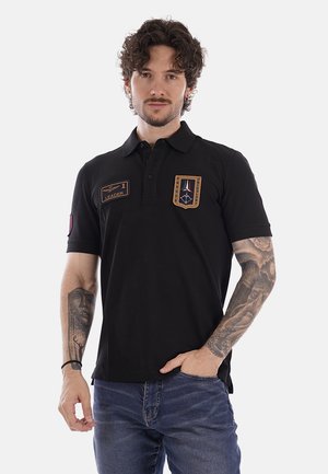 Uomo con capelli ricci che indossa una polo nera con toppe, tatuaggi su entrambe le braccia, in piedi con una mano nella tasca dei jeans.