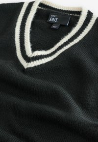 Dunkelgrüner Strickpullover mit V-Ausschnitt und cremefarbenem Streifenbesatz. Weiche Textur, mit einem Etikett in der Nähe des Ausschnitts, das "NEXT EDIT" angibt.