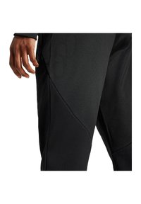 Schwarze Sporthosen mit einem strukturierten Stoff und einer figurbetonten Passform. Die seitlichen Nähte sorgen für eine Formgebung. Eine Hand ruht auf dem Oberschenkel.